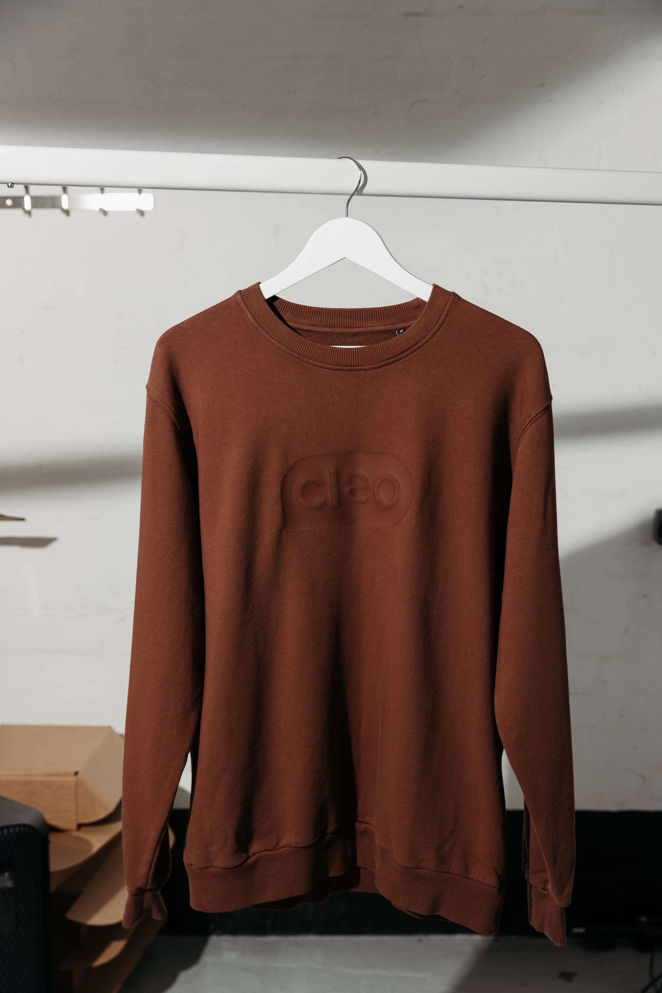 Brown crewneck sweatshirt with a tonal Cleo patch on a white hanger