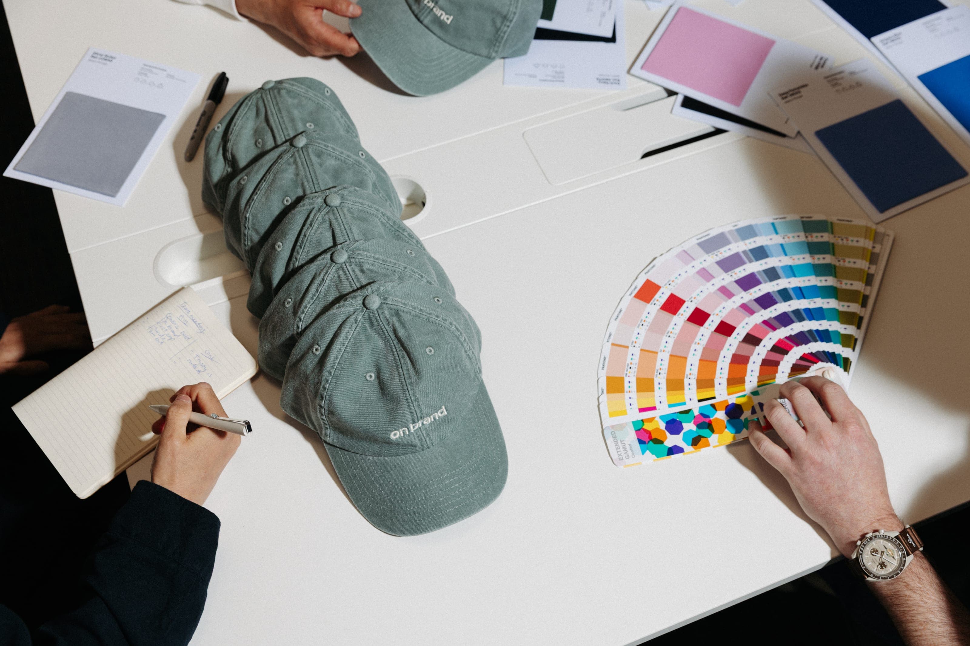 Overhead of stacked sage caps embroidered "on brand" between hands taking notes and holding an open colour fan