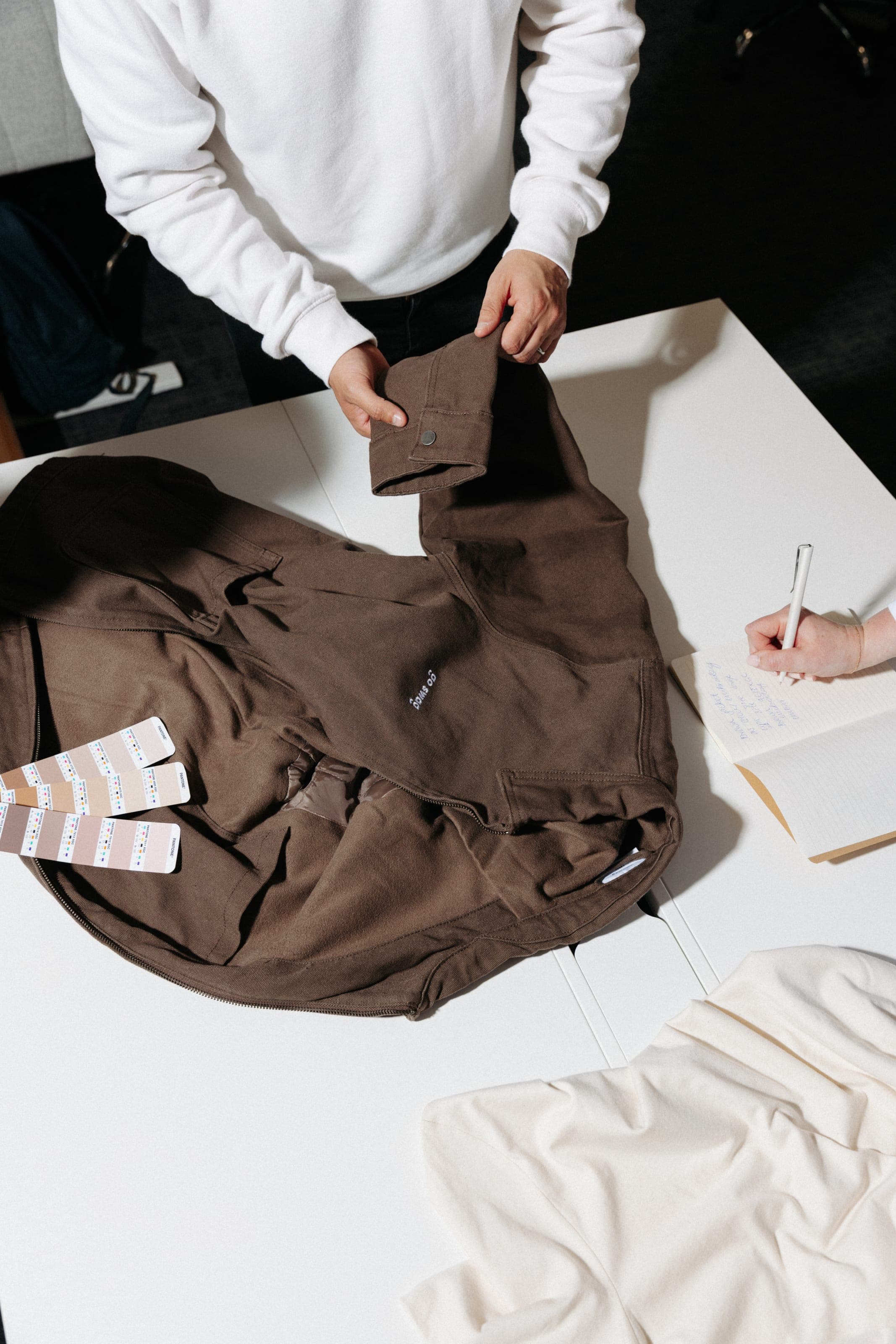 Overhead of hands holding the cuff of a brown overshirt embroidered "go swag" beside brown swatch strips and notes