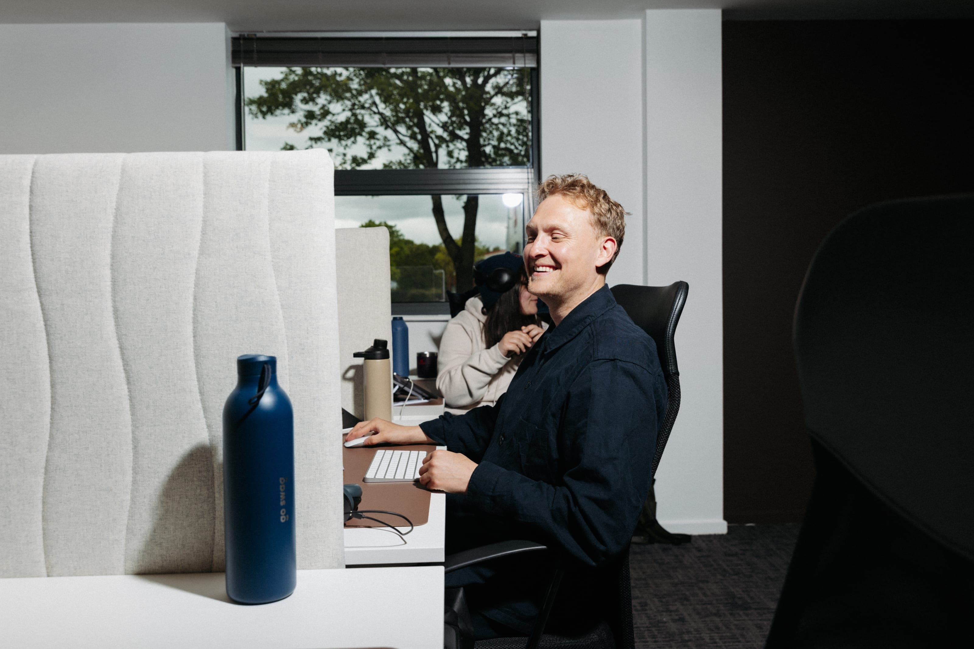 Smiling man at a partitioned desk with a navy go swag bottle in the foreground and a colleague behind