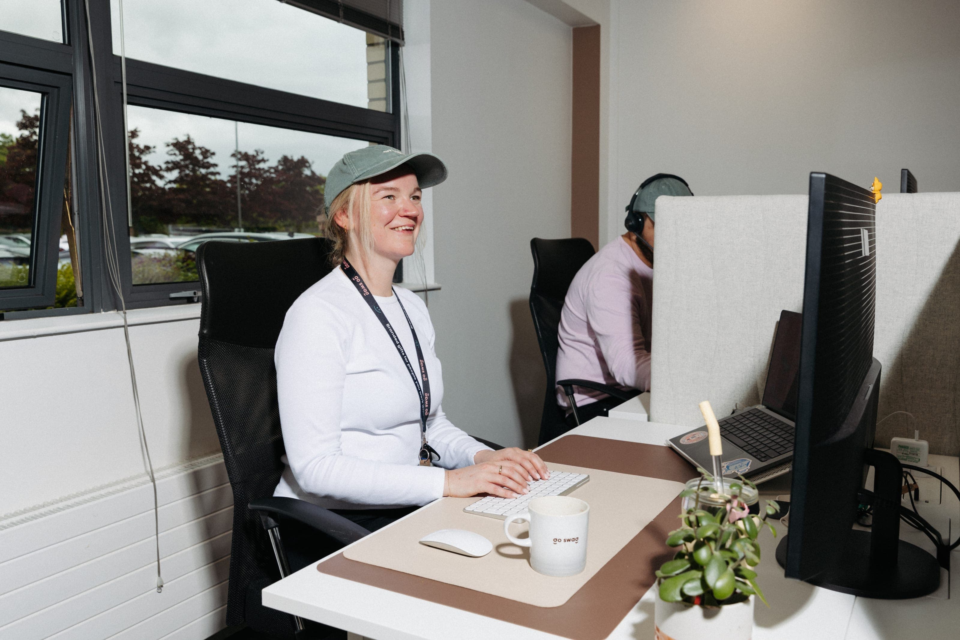 Smiling woman in a sage cap typing at a window desk with a go swag mug, a colleague in a headset behind