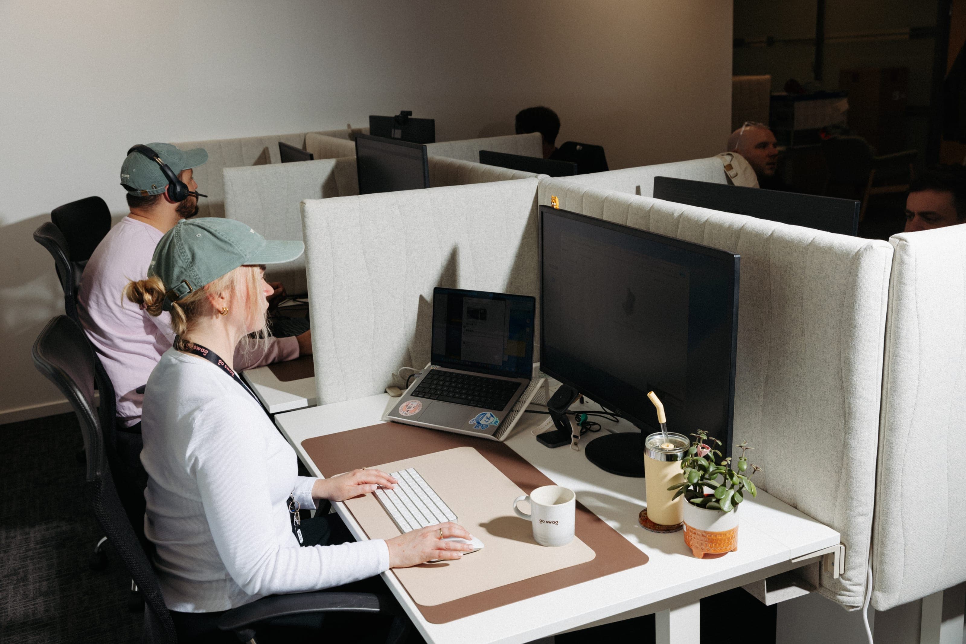 Row of partitioned desks with a woman in a sage cap at a laptop beside a go swag mug and a colleague in a headset