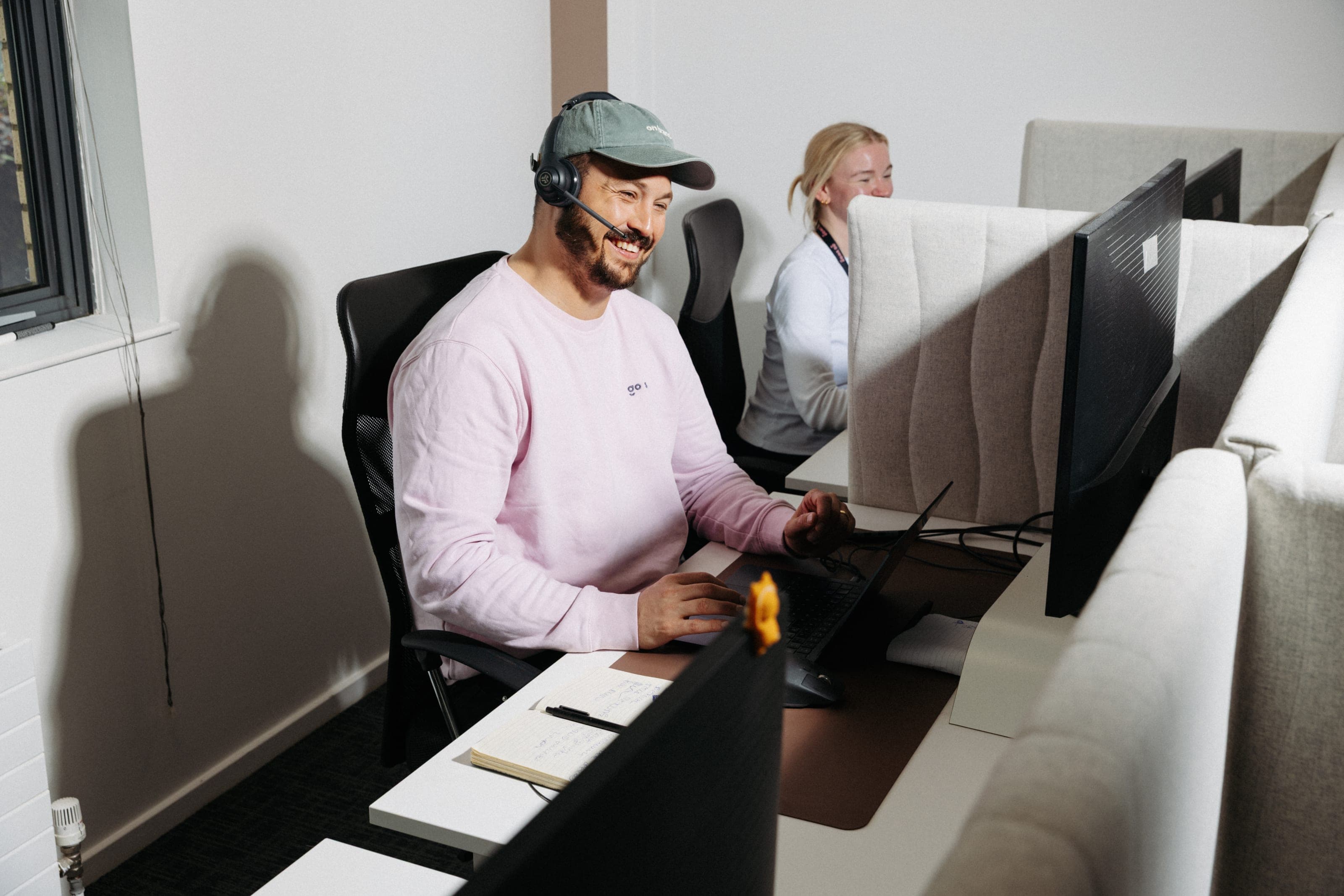 Smiling man wearing a headset and sage cap at his desk, with a colleague working behind him