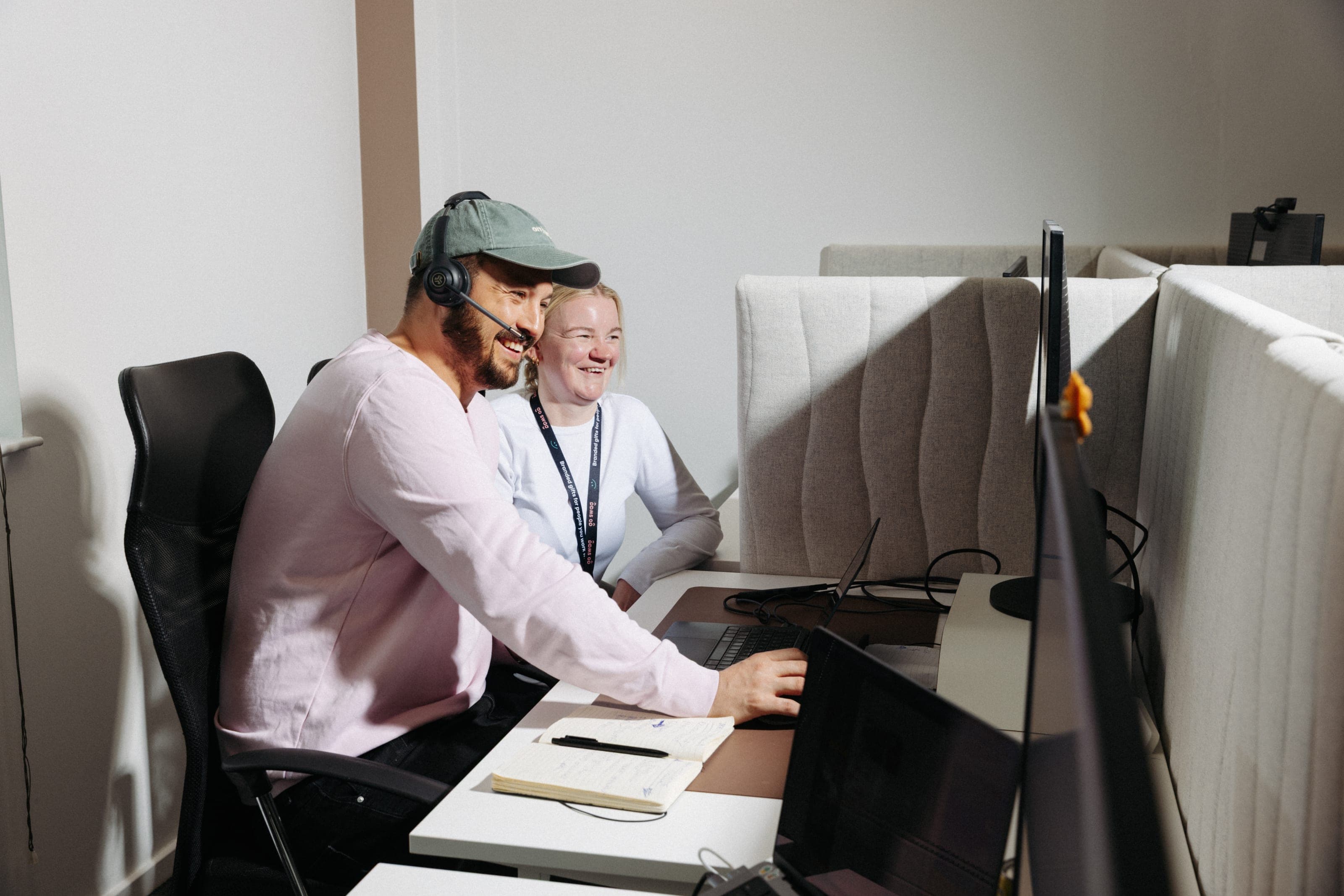 Smiling man in a sage cap and headset and a laughing colleague leaning towards a screen at their desk
