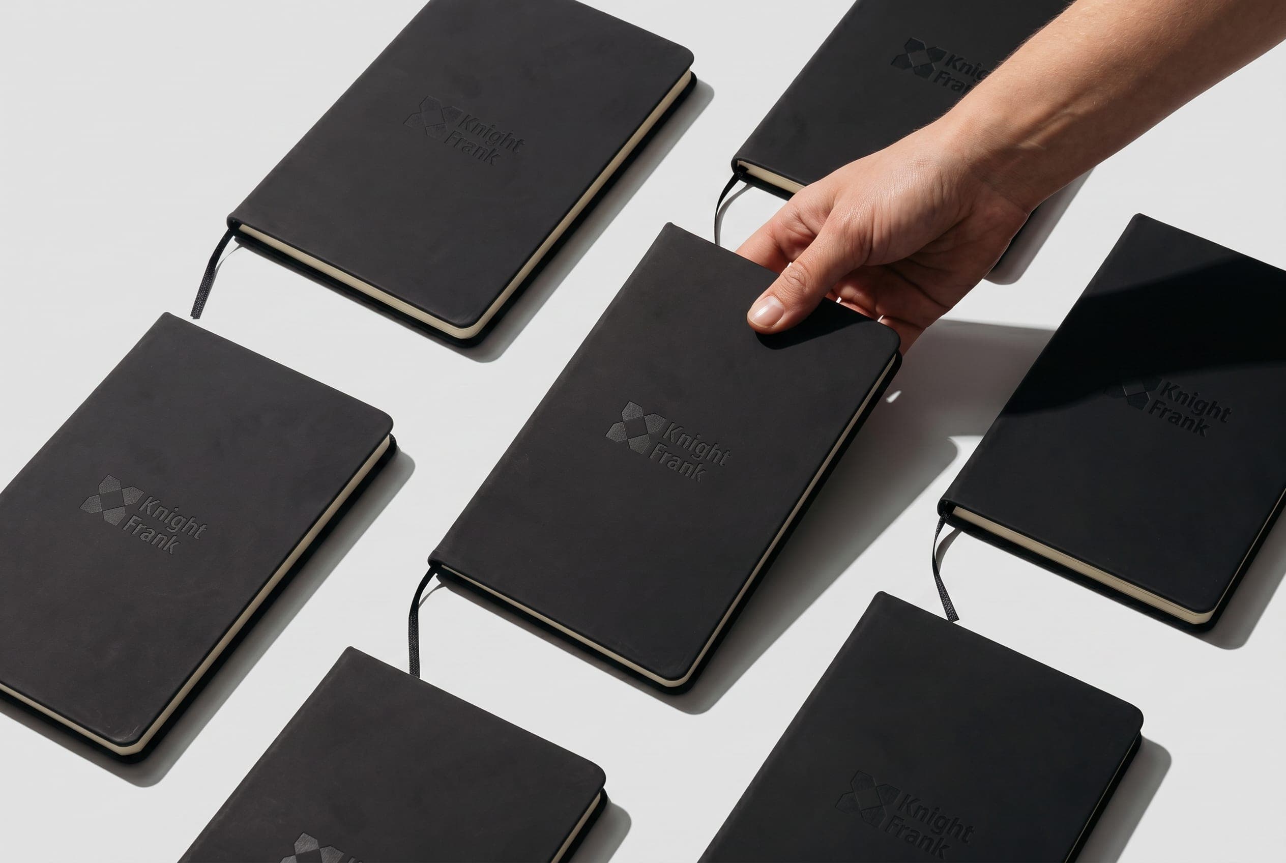 A hand picks up one of several black notebooks debossed with the Knight Frank logo