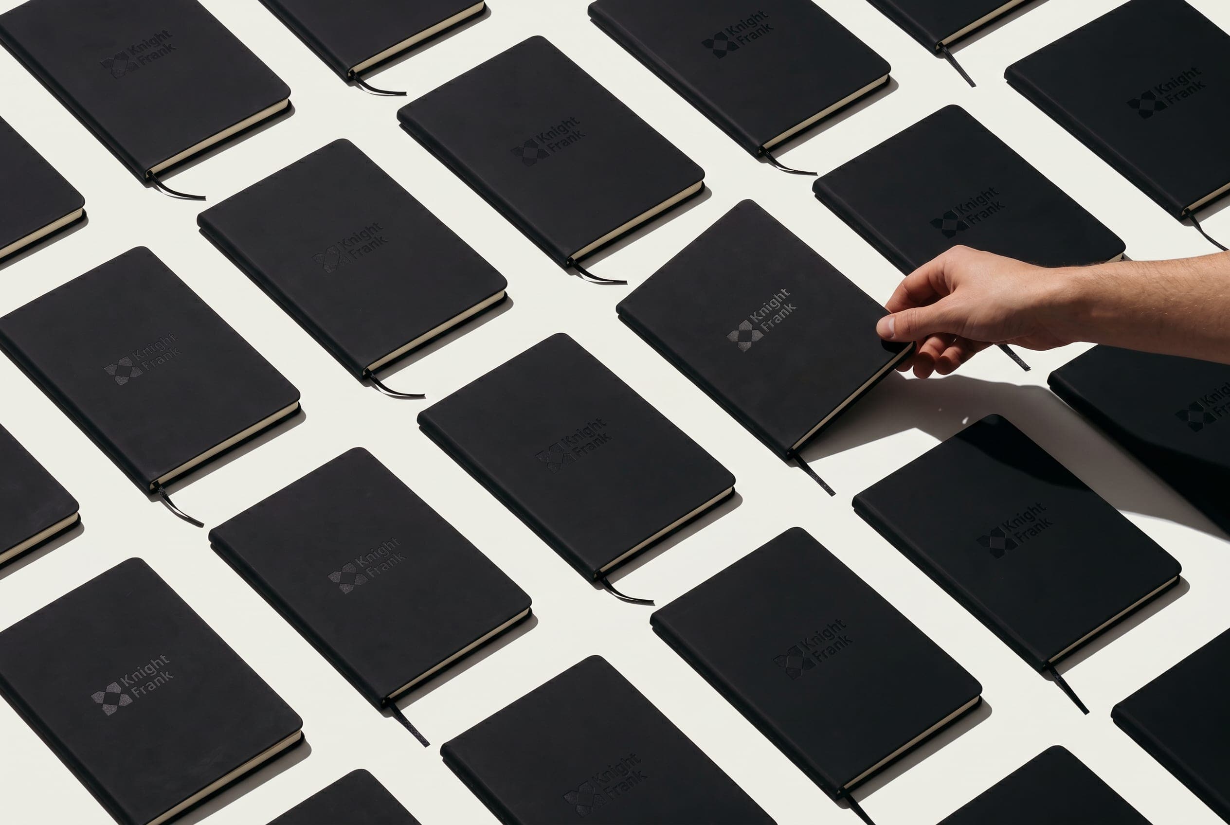 A hand lifts one of dozens of black debossed Knight Frank notebooks laid out in a grid