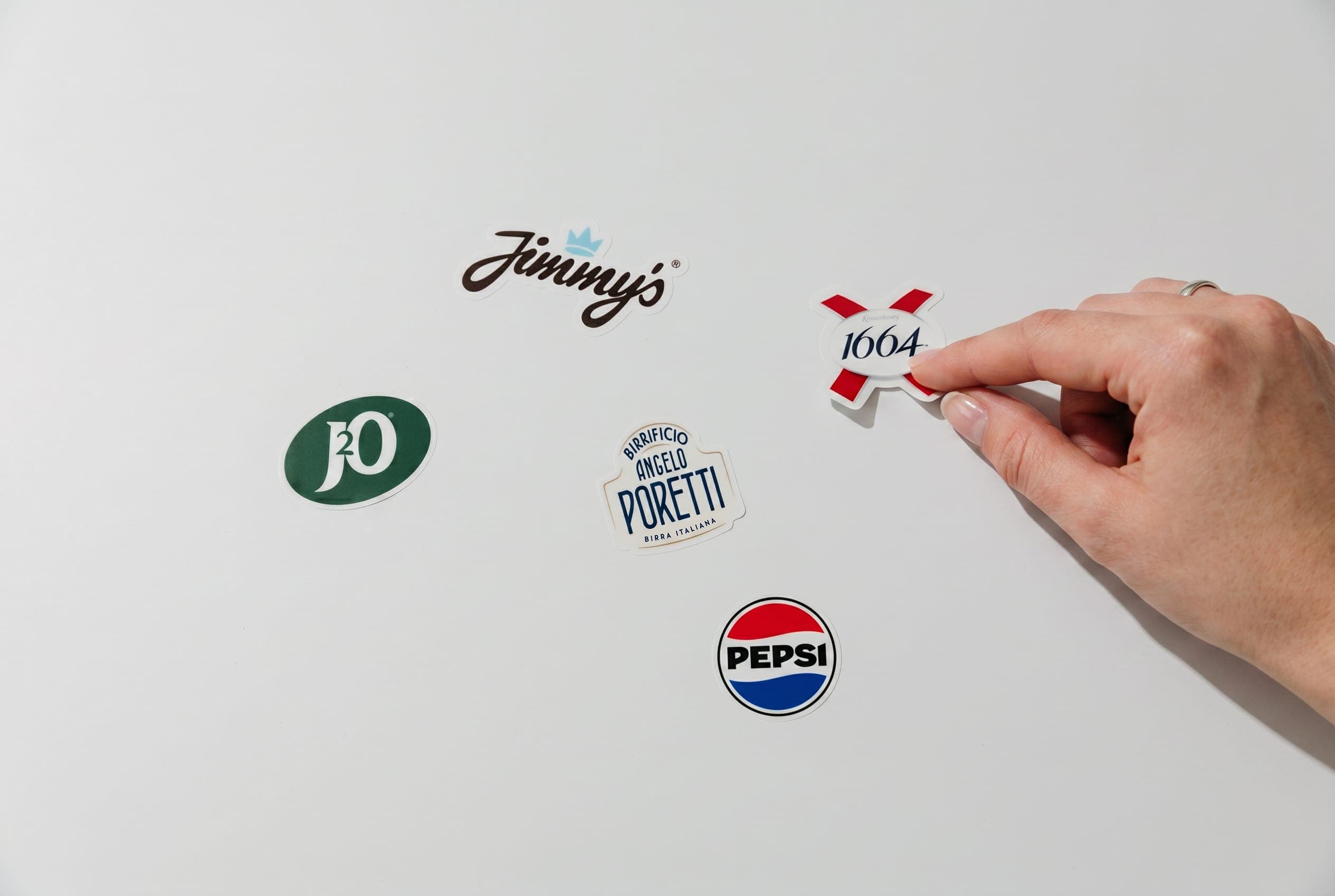A hand places die-cut J2O, Jimmy’s, 1664, Poretti and Pepsi stickers on a white surface
