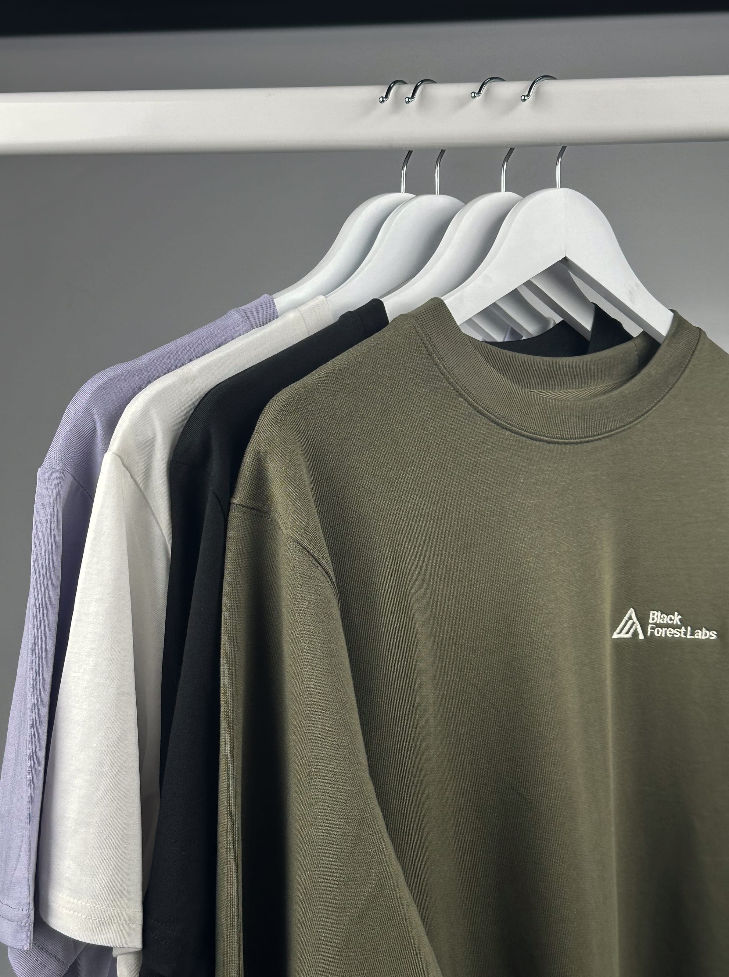 Olive, black, off-white and lilac T-shirts on a rail, the front one embroidered with the Black Forest Labs logo