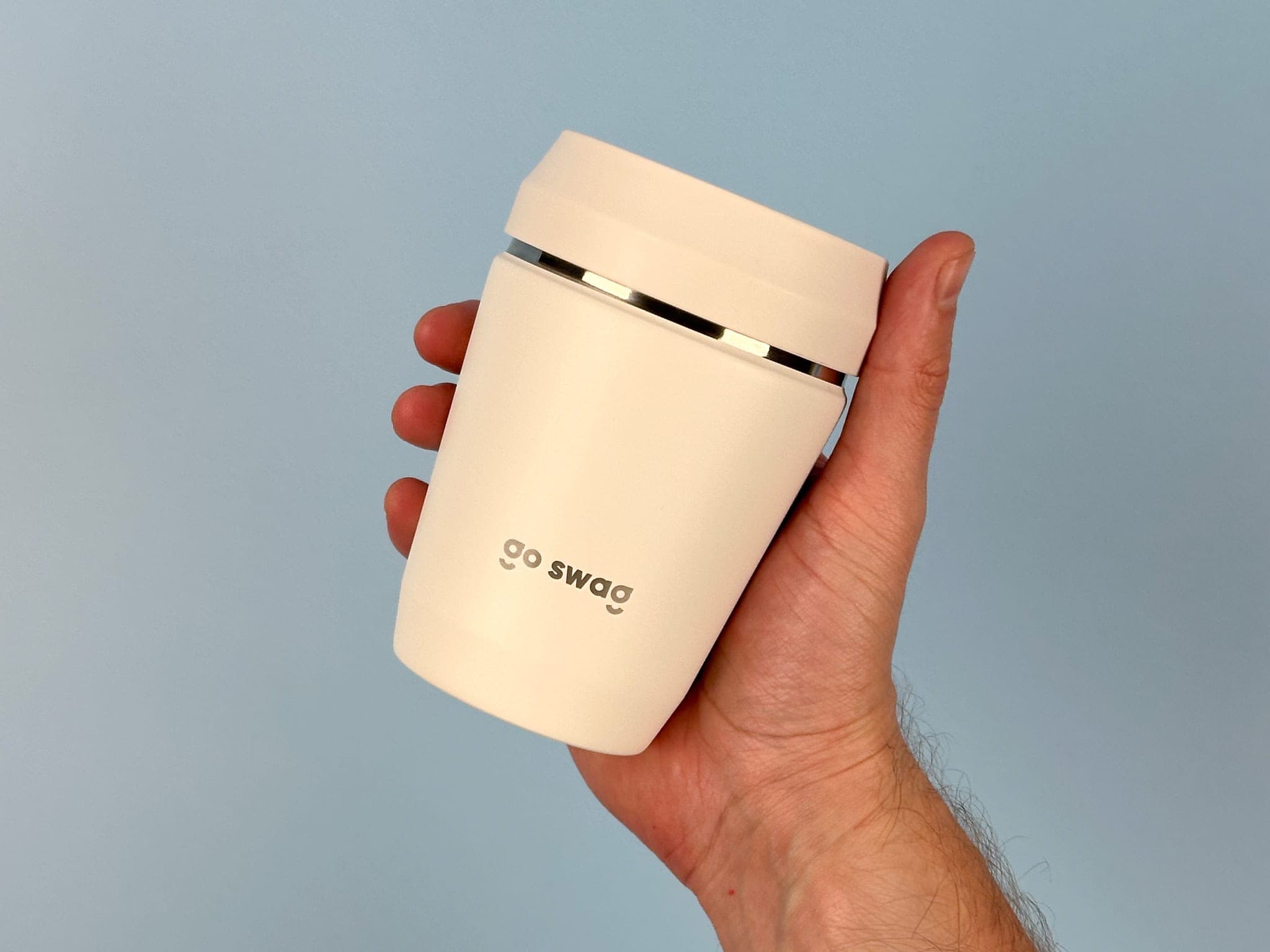 A hand holding up a cream travel cup printed with the grey go swag wordmark against powder blue