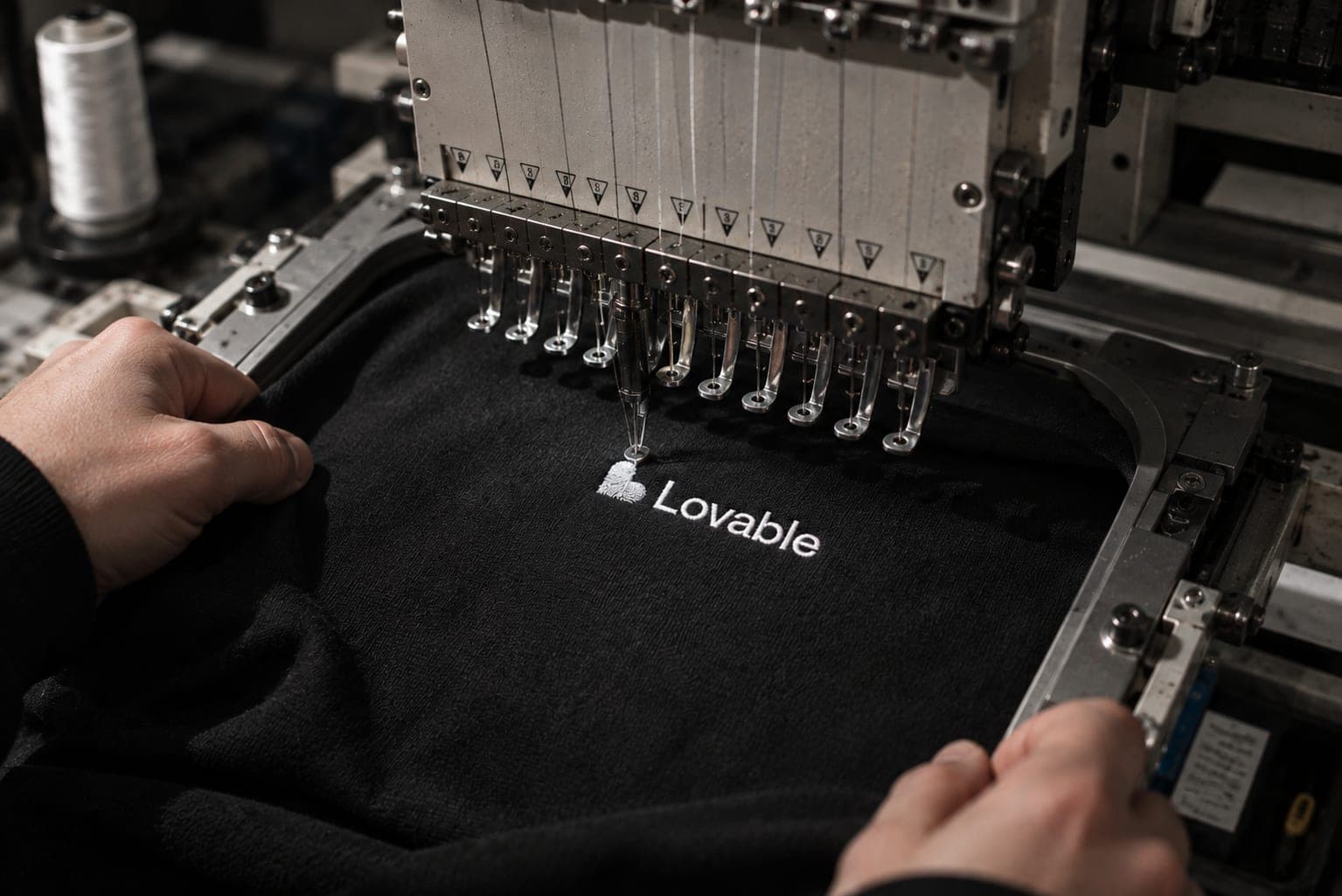 Hands guide a black garment under an embroidery machine as it stitches the Lovable logo in white