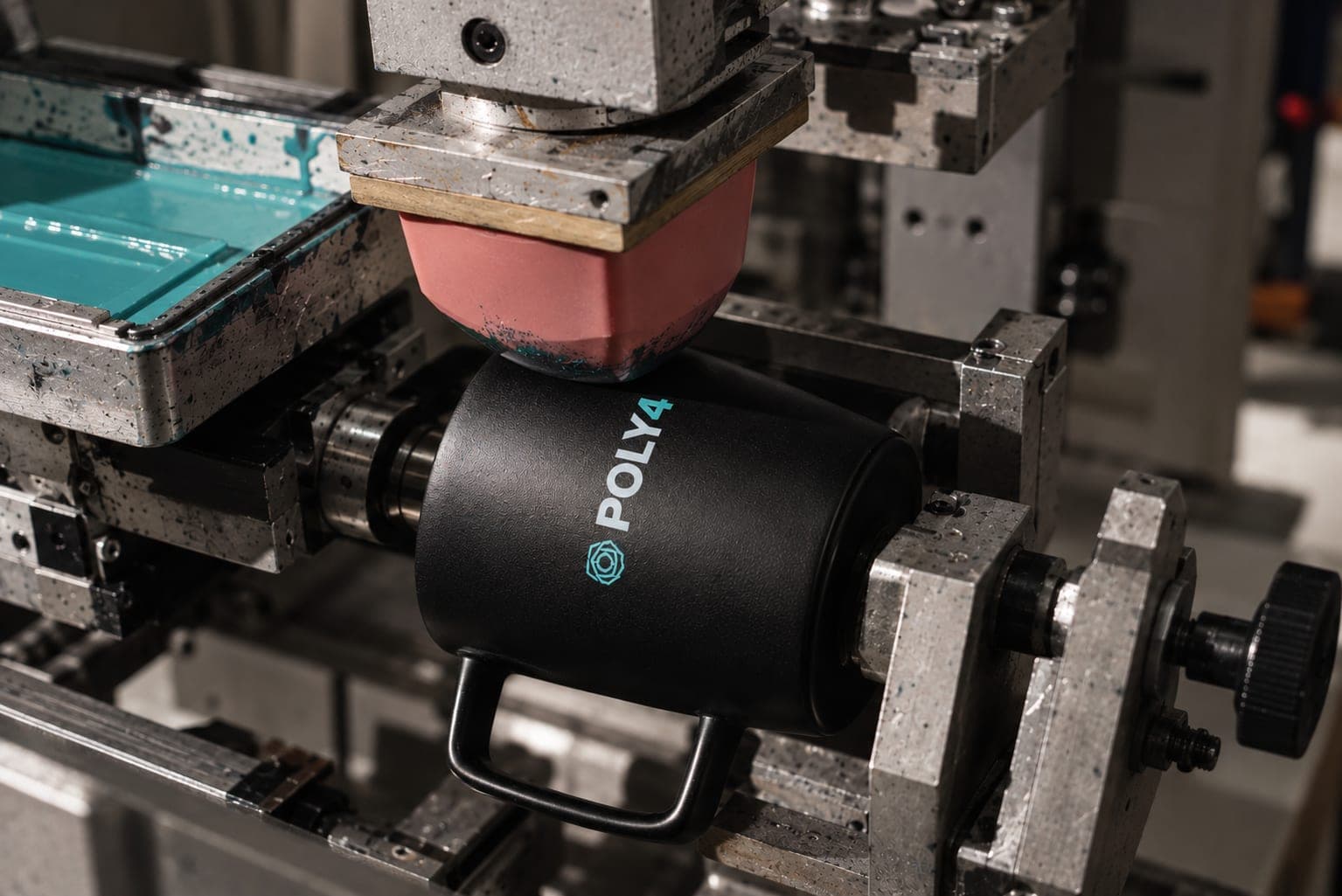 A pink silicone pad presses onto a matte black POLY4 mug in a pad-printing machine, beside a tray of teal ink